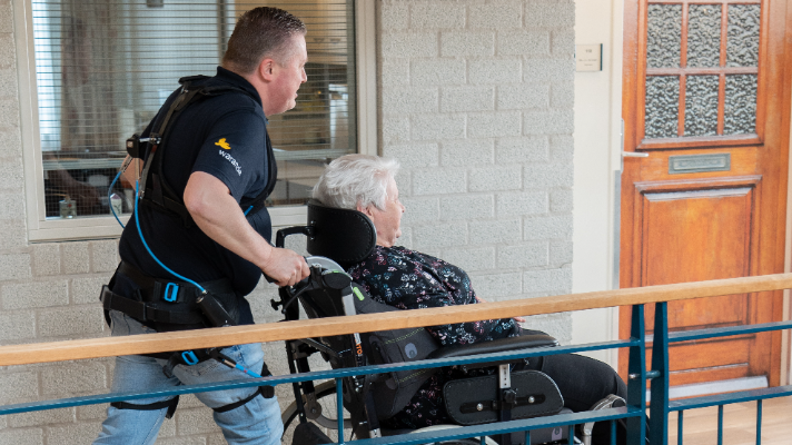 Medewerker met exoskelet aan die bewoner in een rolstoel duwt.